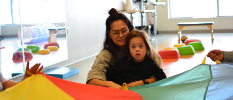 Girl sits on mothers lap in front of rainbow parachute