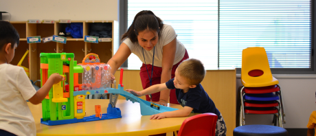 A therapist leans over a table and plays toy cars with a child.