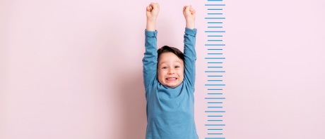Young boy with arms stretched upwards, standing next to measuring lines in front of a pink background.