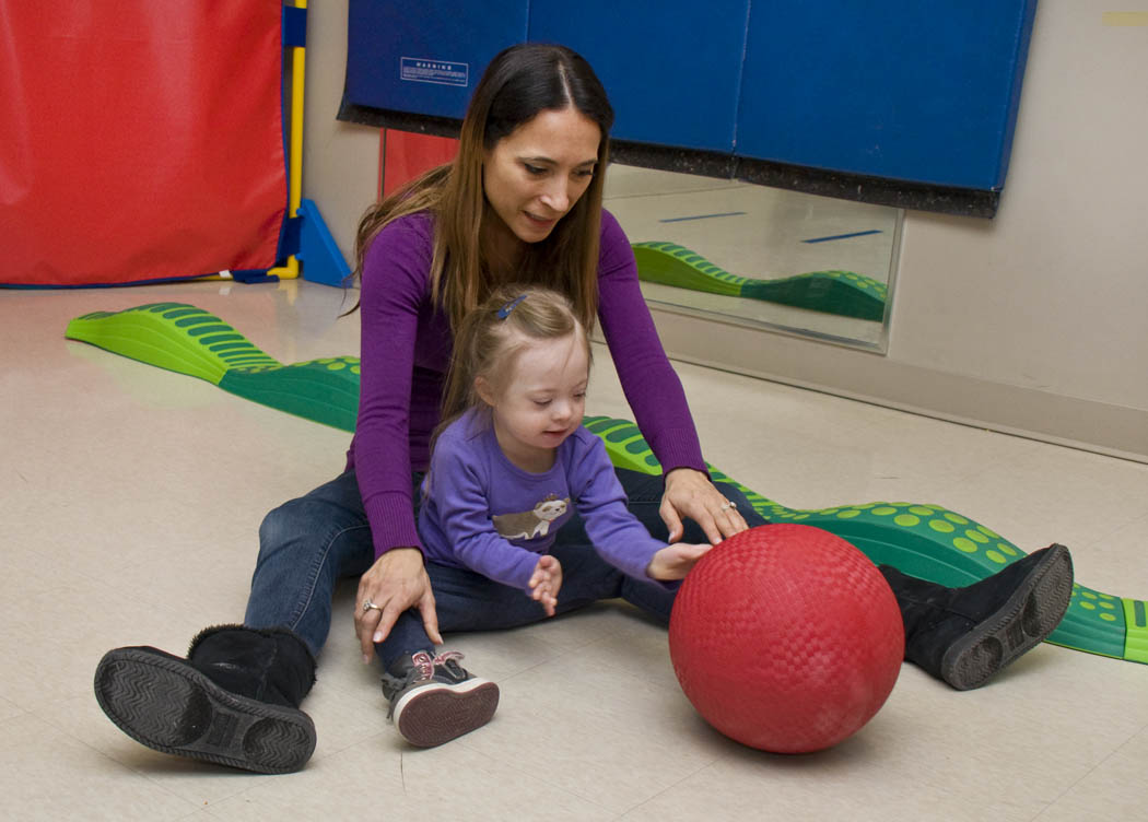 A toddler girl sits on the floor with an adult and catches a red ball rolling towards her