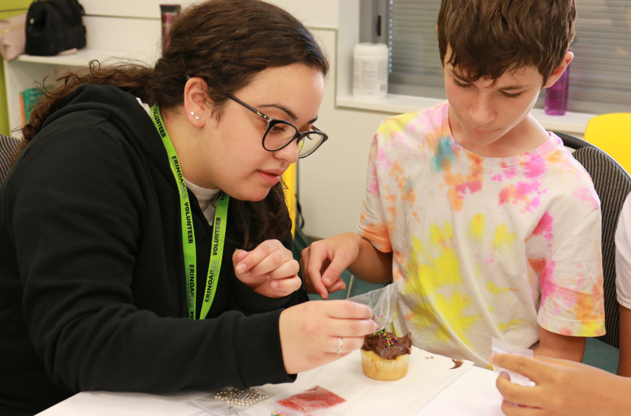 A woman wearing an ErinoakKids lanyard assists a young boy in decorating a cupcake