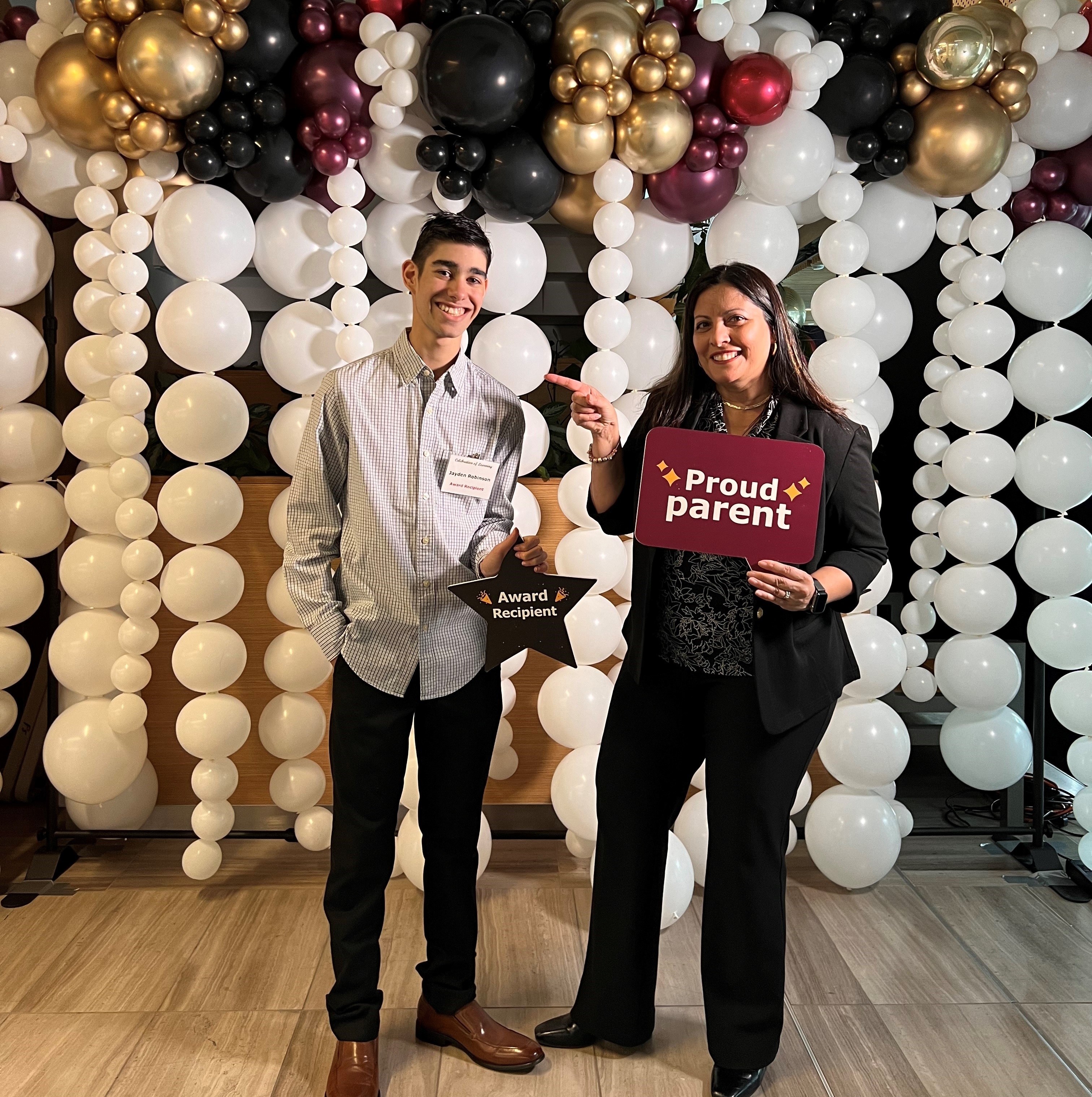 Jayden holding a sign that reads "Award recipient" with his mother who is holding a sign that reads "proud parent". There are balloons in the background.